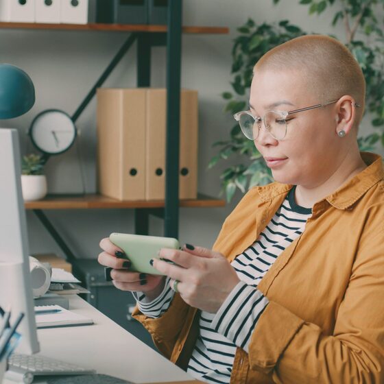 A woman with a shaved head sitting at a desk at home, holding a tablet.
