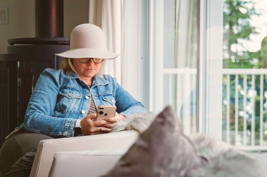 A woman in a wide brimmed hat sitting by a window at home, looking at her phone.